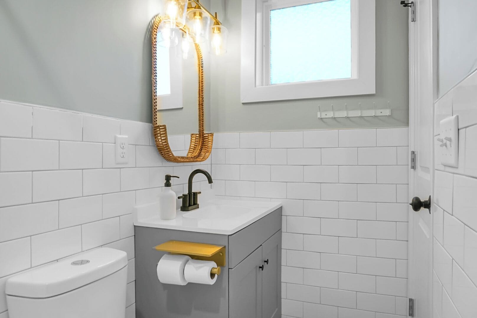 Une salle de bain avec des murs recouverts de carreaux blancs, un lavabo blanc avec un robinet doré, des toilettes et une fenêtre située au-dessus du lavabo.