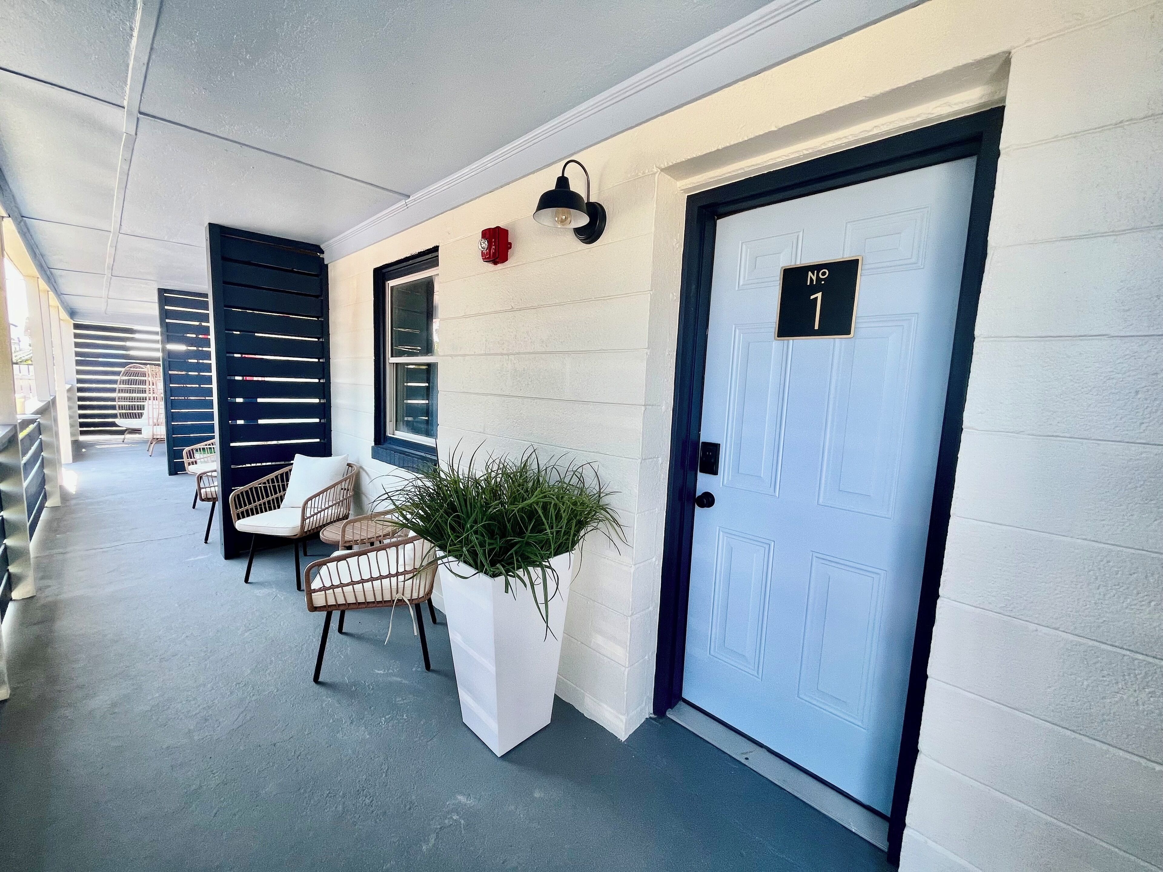 A covered outdoor seating area with chairs and a table, a white door with a number plaque, and a wall-mounted light fixture.