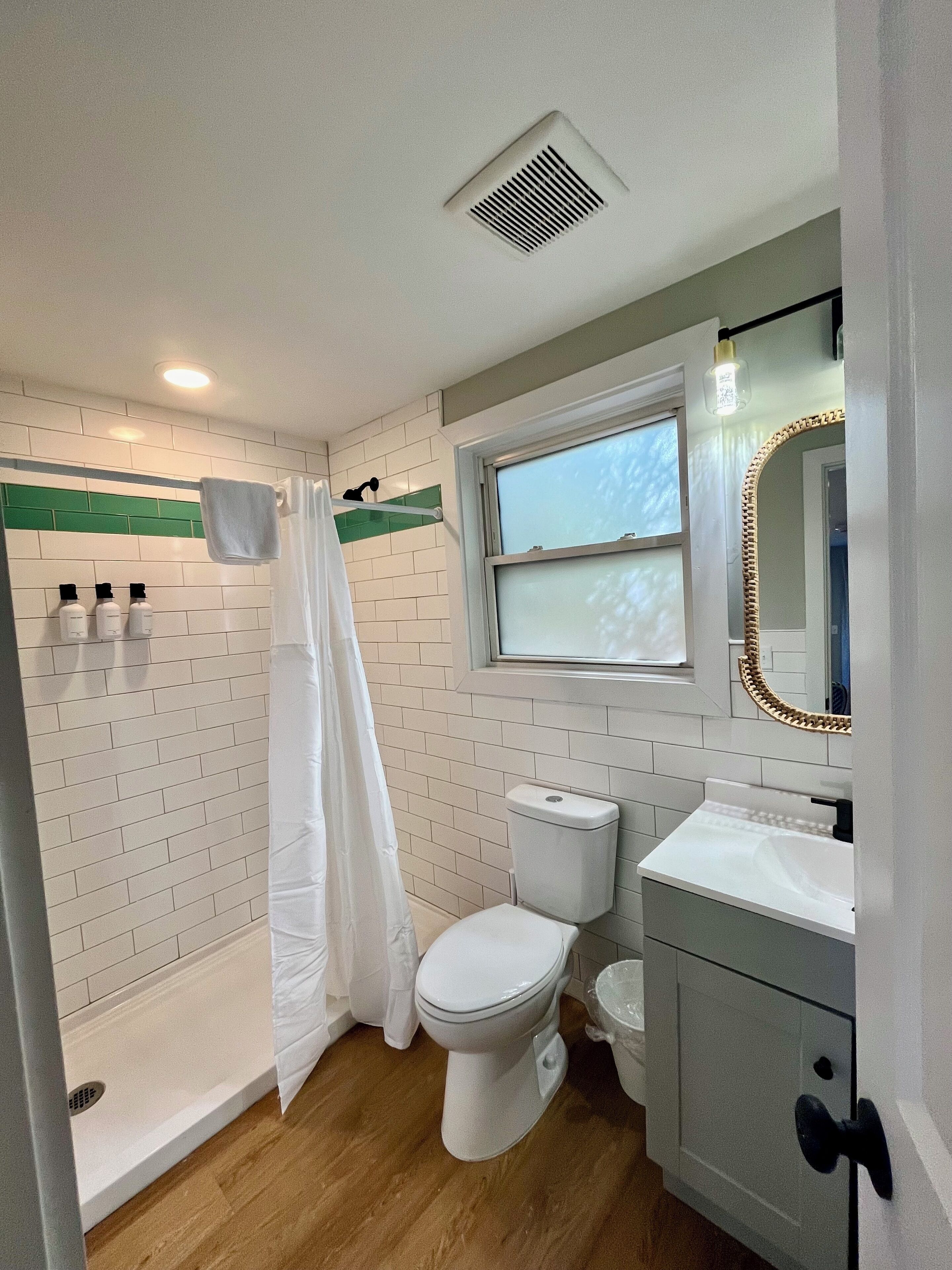 Un baño con inodoro blanco, ducha con cortina blanca, lavamanos con espejo y una ventana sobre el lavamanos.