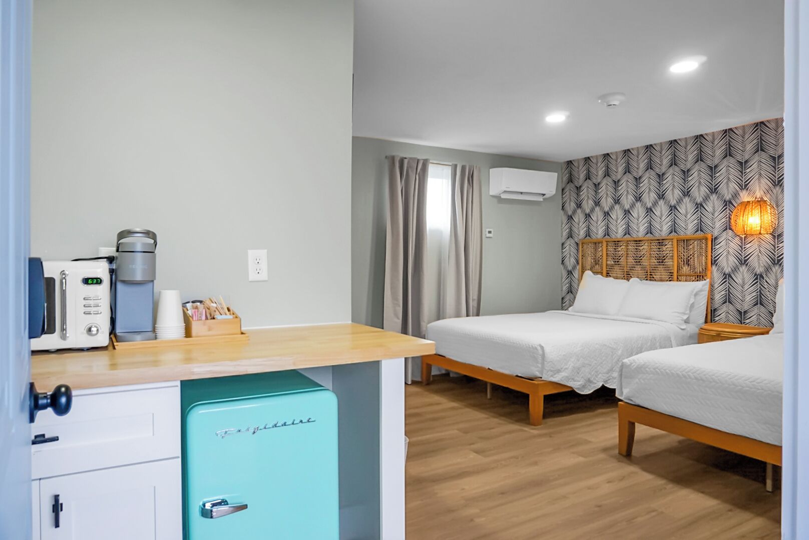 A compact hotel room with a kitchenette, two single beds, a small fridge, and a patterned wall.