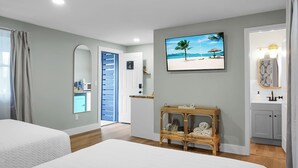 Een hotelkamer met twee bedden, een badkamer met spiegel en wastafel, een zithoekje met een rieten tafeltje, en een schilderij met een strandthema aan de muur.