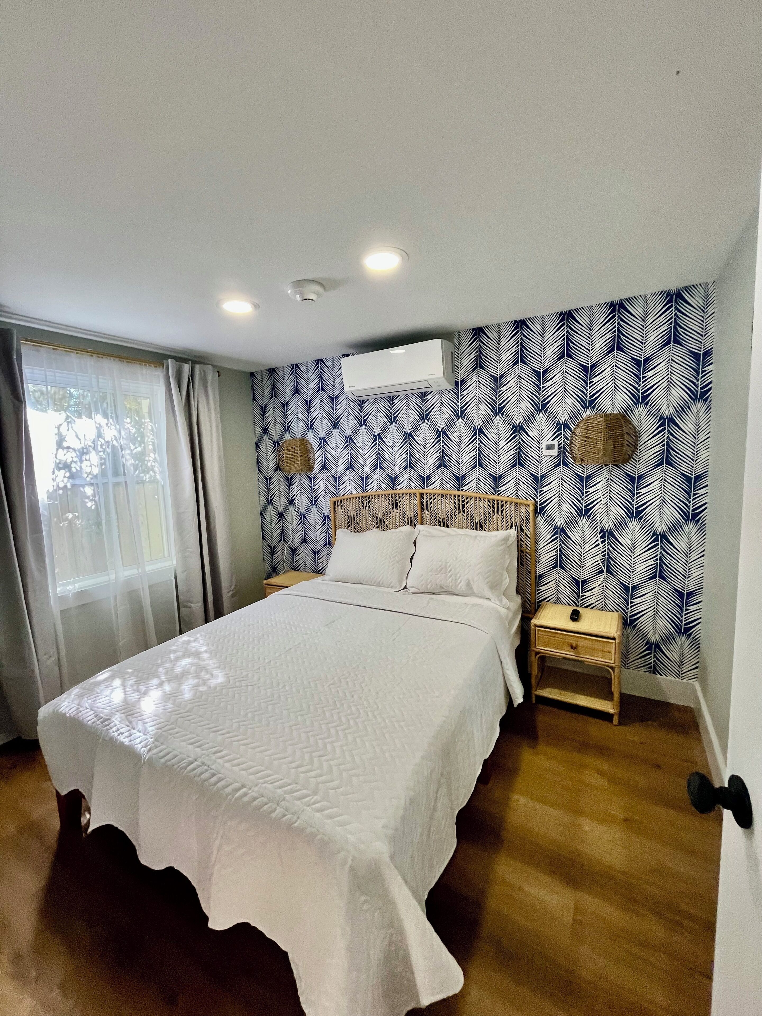 A bedroom with a bed, a nightstand, a wall-mounted air conditioner, and a window with curtains.