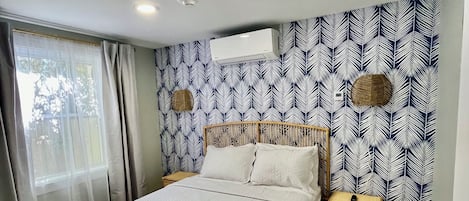 A bedroom with a bed, a nightstand, a wall-mounted air conditioner, and a window with curtains.
