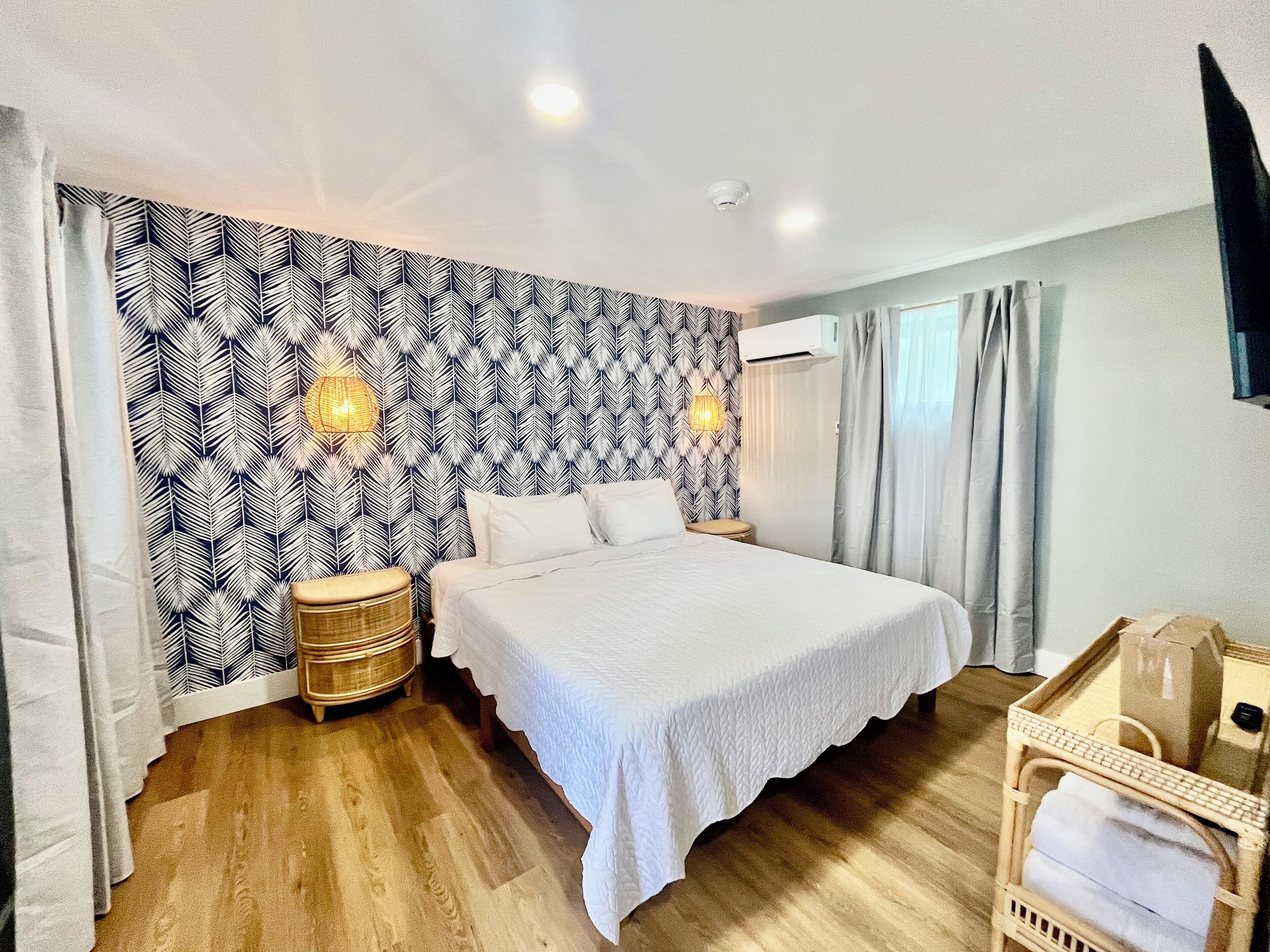 Un dormitorio con papel tapiz estampado, una cama con ropa de cama blanca, dos lámparas de pared, una mesita de mimbre y un suelo de madera.
