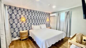Un dormitorio con papel tapiz estampado, una cama con ropa de cama blanca, dos lámparas de pared, una mesita de mimbre y un suelo de madera.