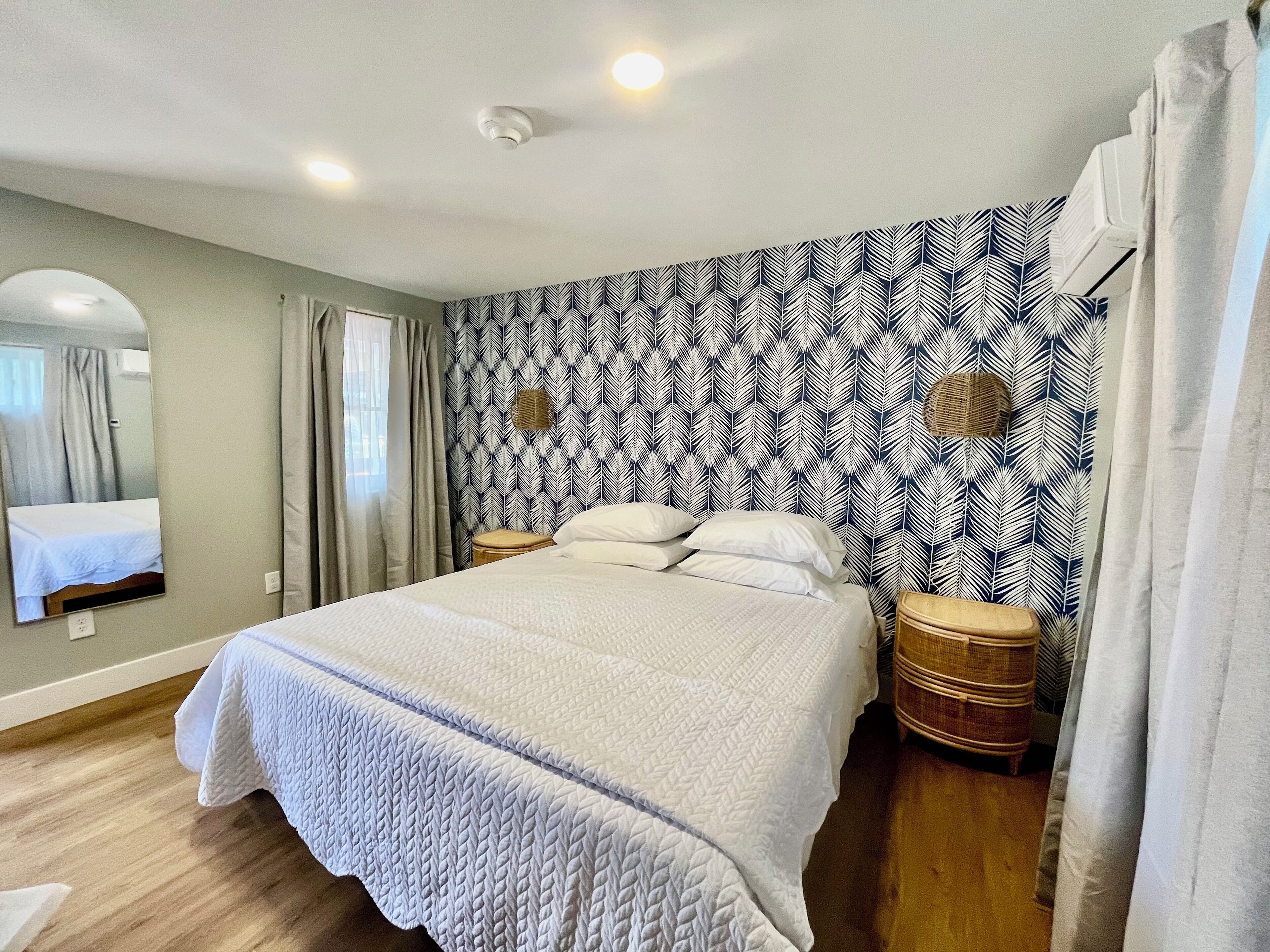 A bedroom with a bed, a mirror, a window with curtains, a patterned wall, and a ceiling light.