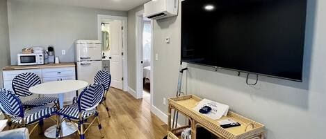 A modern living room with a dining area, a flat-screen TV mounted on the wall, a kitchen in the background, and a wicker side table with towels.