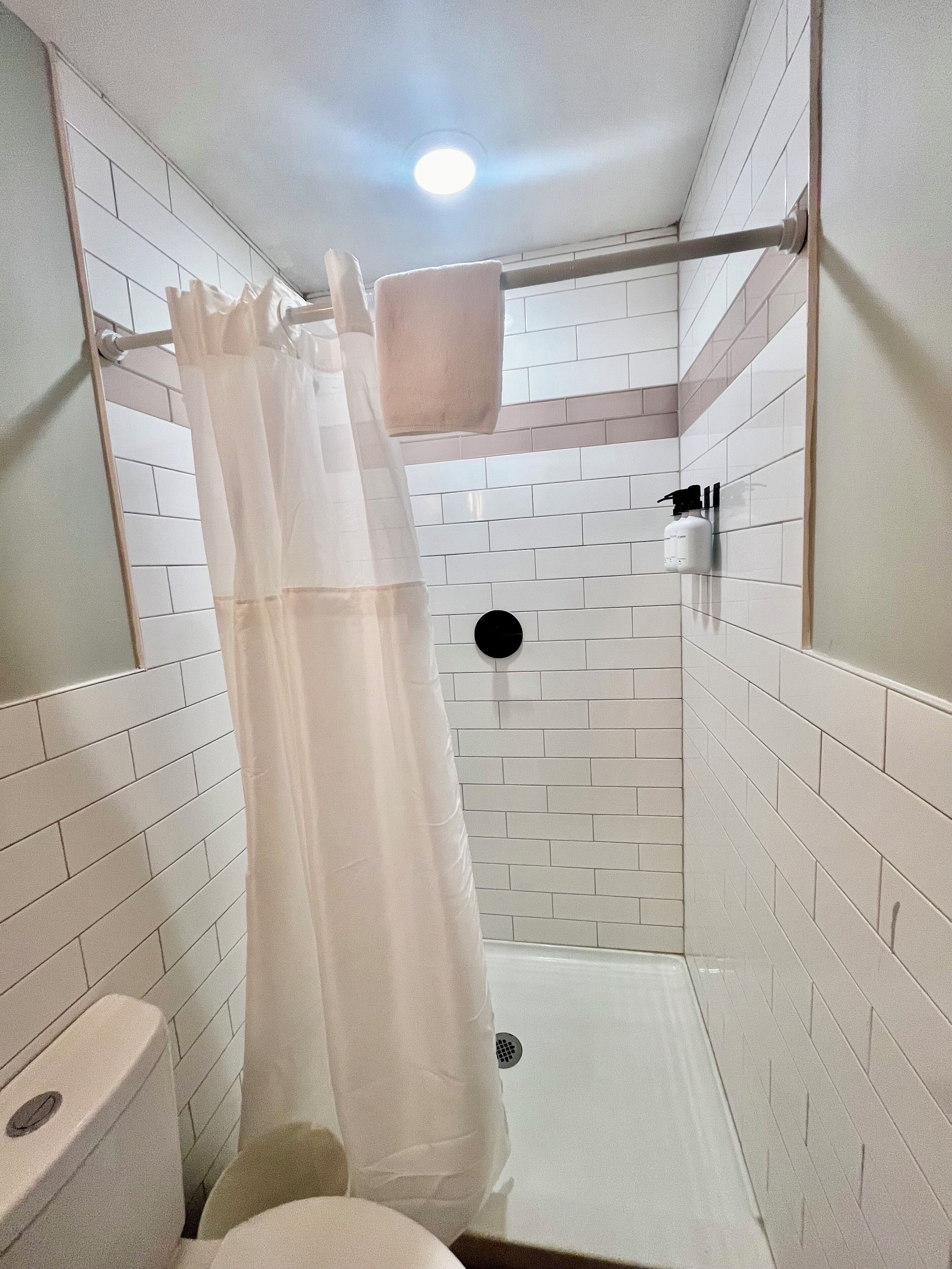 Un baño pequeño con azulejos blancos, cortina de ducha, inodoro y dispensador de jabón en la pared.