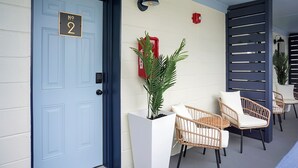 Een blauwe deur met een cijferbord, een potplant, rieten stoelen en een wandlamp.