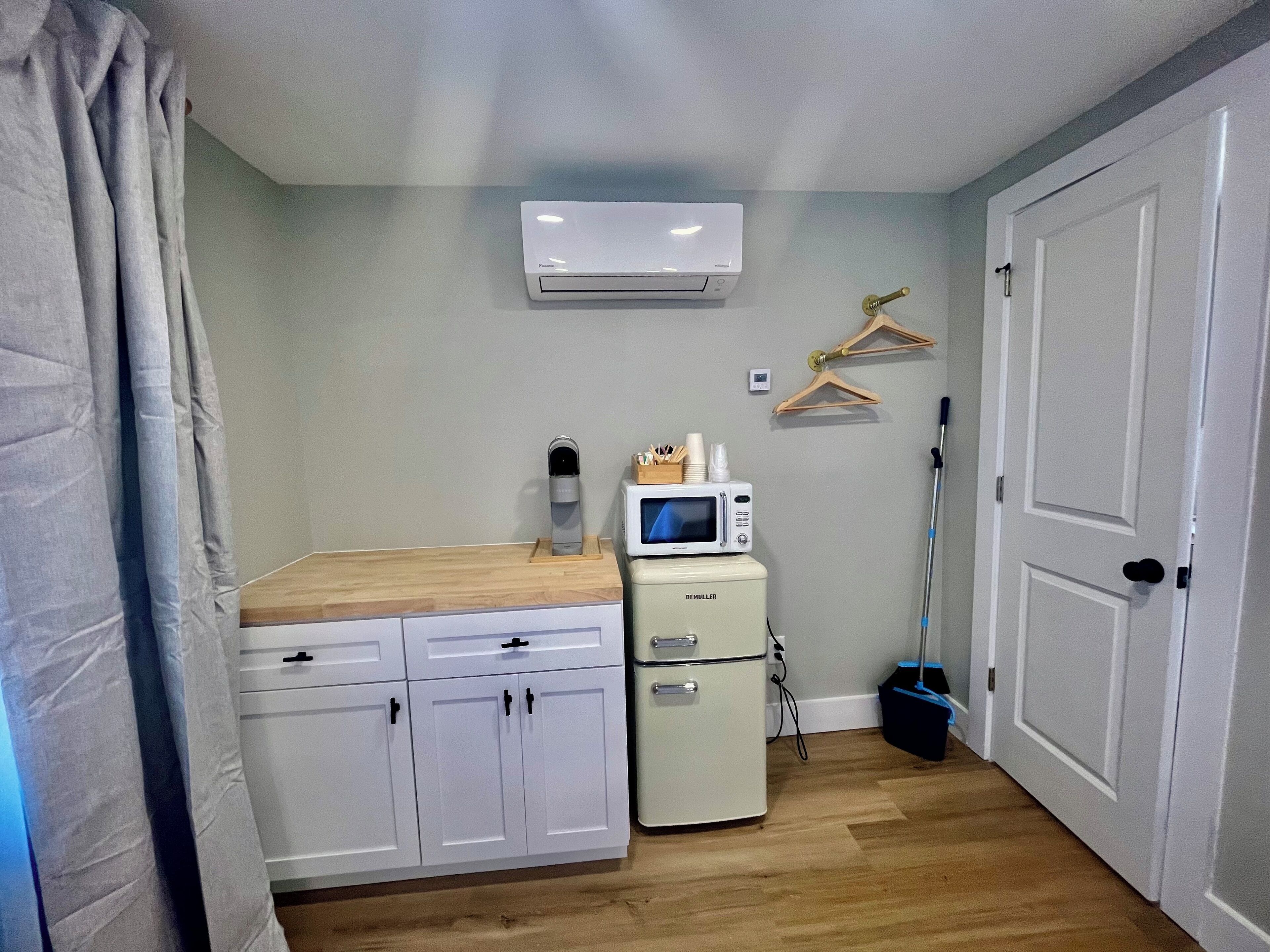 Ein gemütlicher Raum mit einer Küchenzeile, einem weißen Kühlschrank, einer Mikrowelle, einem kleinen Schrank, einer Holz Arbeitsplatte, einer Decken-Klimaanlage, einem Besen und einer Tür.