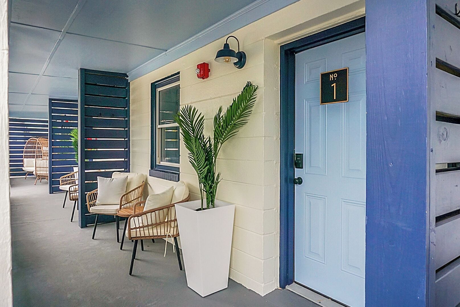 Un espacio de estar al aire libre cubierto con sillas de mimbre y una planta en maceta, una puerta azul con placa de número y un aplique de pared.
