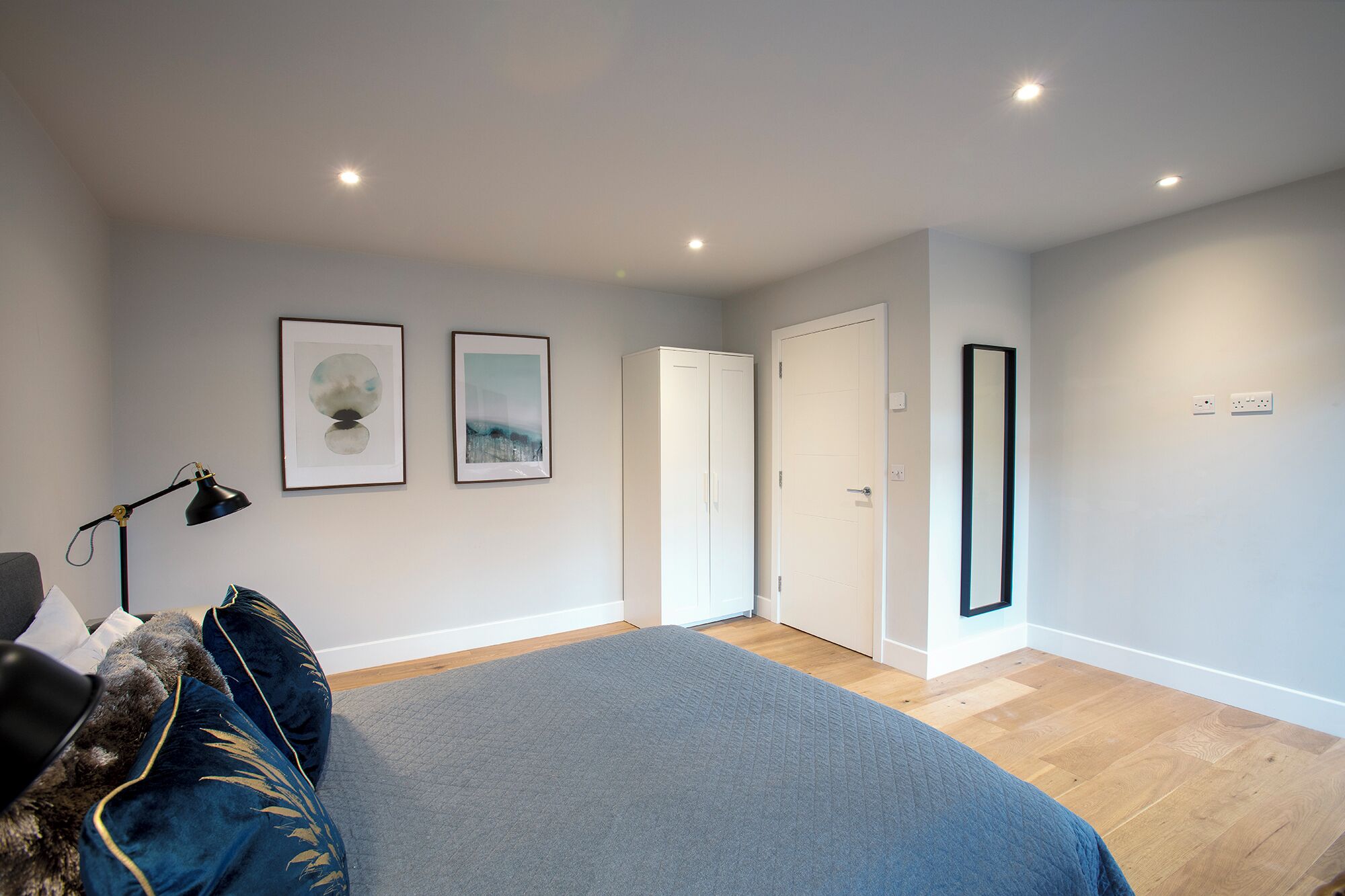 A bedroom with a bed, a desk, a chair, a lamp, and two framed artworks on the wall.