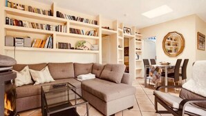 Ein Wohnzimmer mit Ecksofa, Glastisch und Kamin. Bücherregale voller Bücher und ein Essbereich mit Tisch und Stühlen sind ebenfalls vorhanden.