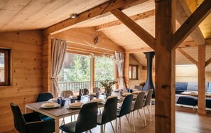 Una cabaña de madera con comedor que incluye una mesa dispuesta para seis comensales, sillas y un amplio ventanal con cortinas.