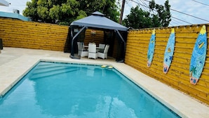 A rectangular swimming pool with a covered seating area and surfboard decorations on the wall.