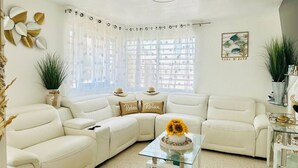 A living room with a sectional sofa, a glass coffee table with a sunflower centerpiece, and a large window with sheer curtains.
