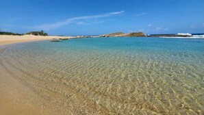Ein Strand mit klarem, seichten Wasser, Sandküste und in der Ferne felsigen Felsen unter blauem Himmel.