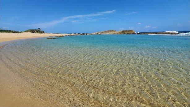 A beach with clear shallow waters, sandy shore, and distant rocky outcrops under a clear blue sky.