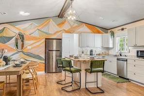 A modern kitchen with a mountain mural, wooden dining table, and green bar stools.