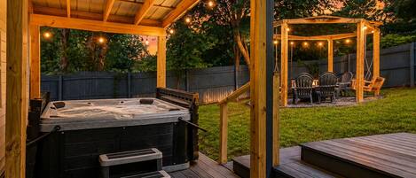 Un jacuzzi bajo una pérgola de madera con luces colgantes, una terraza y un pabellón en un patio trasero.