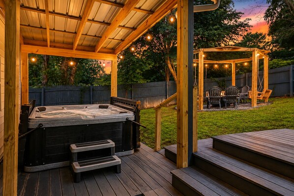 A hot tub under a wooden pergola with string lights, a deck, and a gazebo in a backyard.