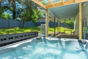 Ein Whirlpool unter einer Holzpergola in einem Garten mit Zaun, Grill und Stühlen.