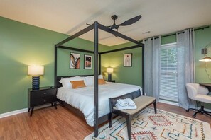 A four-poster bed with a ceiling fan, a bedside table with a lamp, a chair, and a patterned rug.