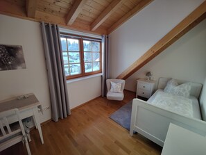 Un dormitorio con techo inclinado, una cama, una mesita de noche, una silla, un escritorio y una ventana con cortinas.