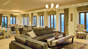 A spacious living area with a sofa, coffee table, and a colorful armchair. There are multiple windows providing natural light and a view of the outdoors. - Arktos hotel (Ioannina)