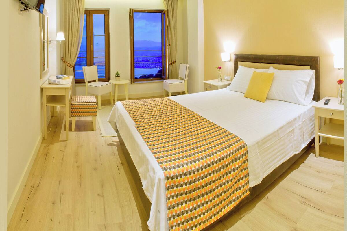 Habitación de hotel con una cama grande, un escritorio, una silla y vistas al mar a través de dos ventanas.