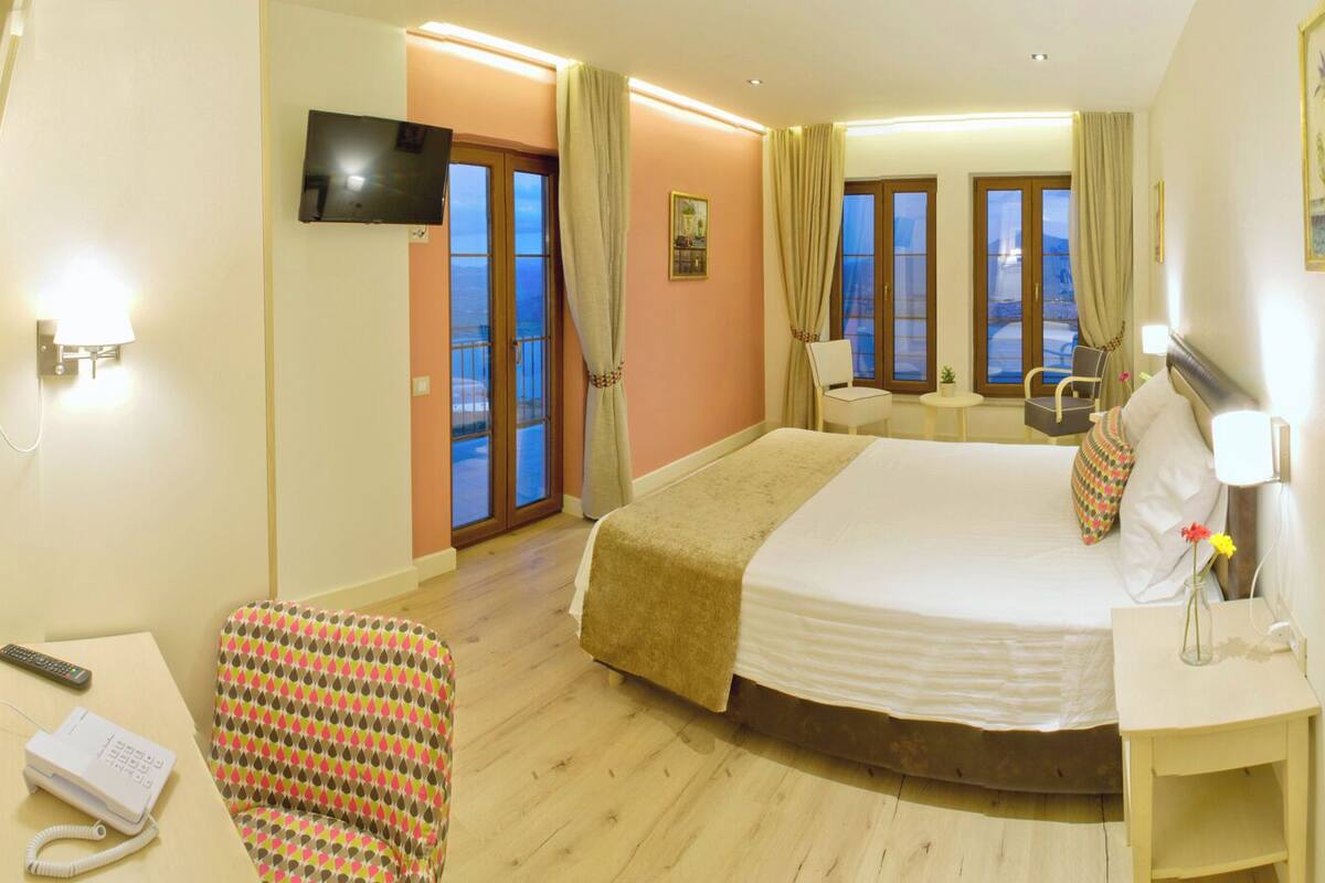 Habitación de hotel con cama, escritorio, silla, televisor y balcón con cortinas.