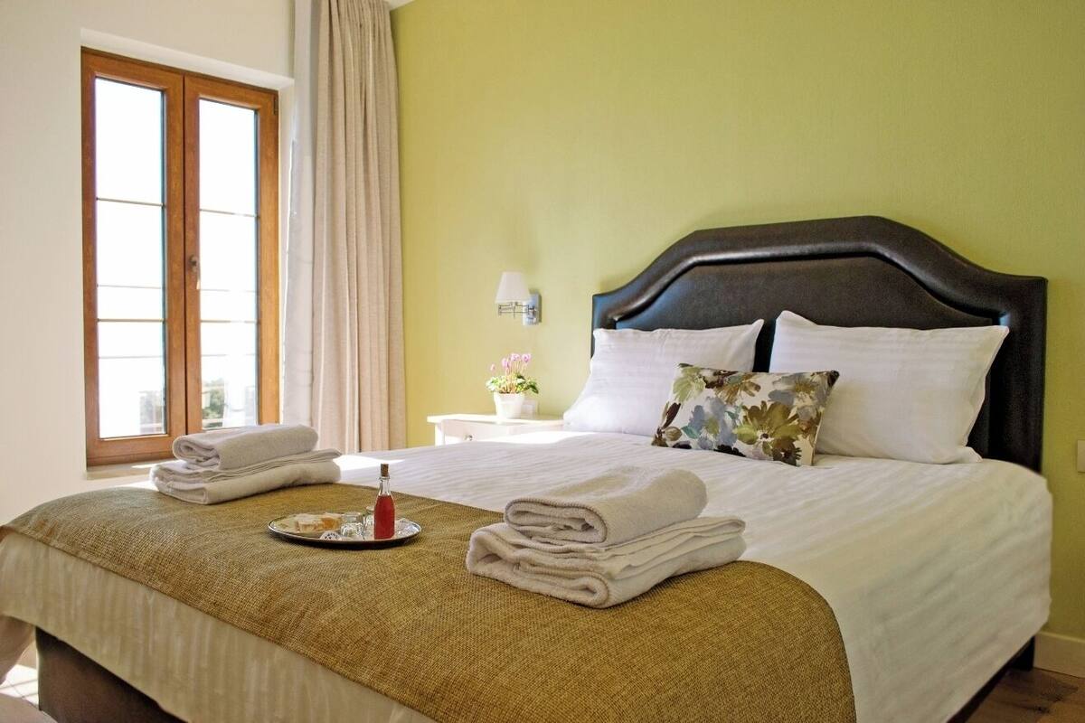 Una cama bien hecha con sábanas blancas, una mesita de noche con lámpara y una ventana con cortinas.