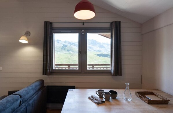 A room with a wooden wall, a window with curtains, a table with cups and a pitcher, and a bench.