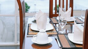 A dining table set with white plates, napkins, and a white teapot, with a view of the ocean and a blue umbrella in the background. - Surendra Seaview (Matara)