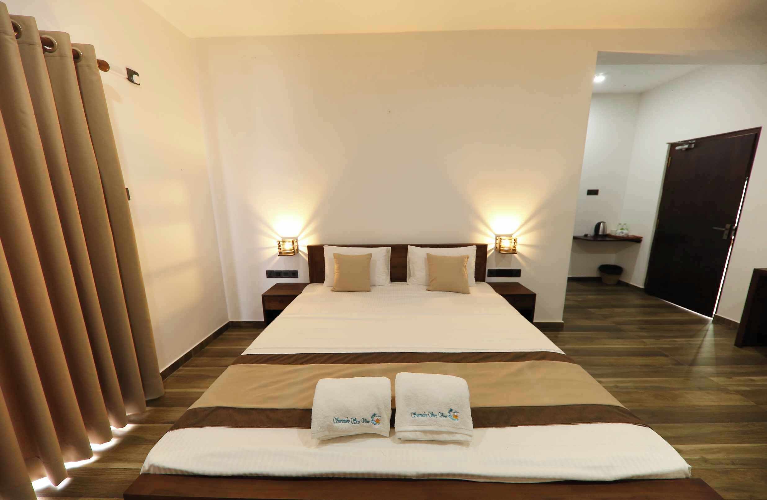 Habitación de hotel con una cama grande, dos mesitas de noche con lámparas, piso de madera y puerta al baño.