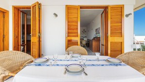 Un espace extérieur aménagé pour les repas, avec une table dressée pour quatre personnes, entourée de chaises en osier, et des portes en bois donnant accès à un espace intérieur.
