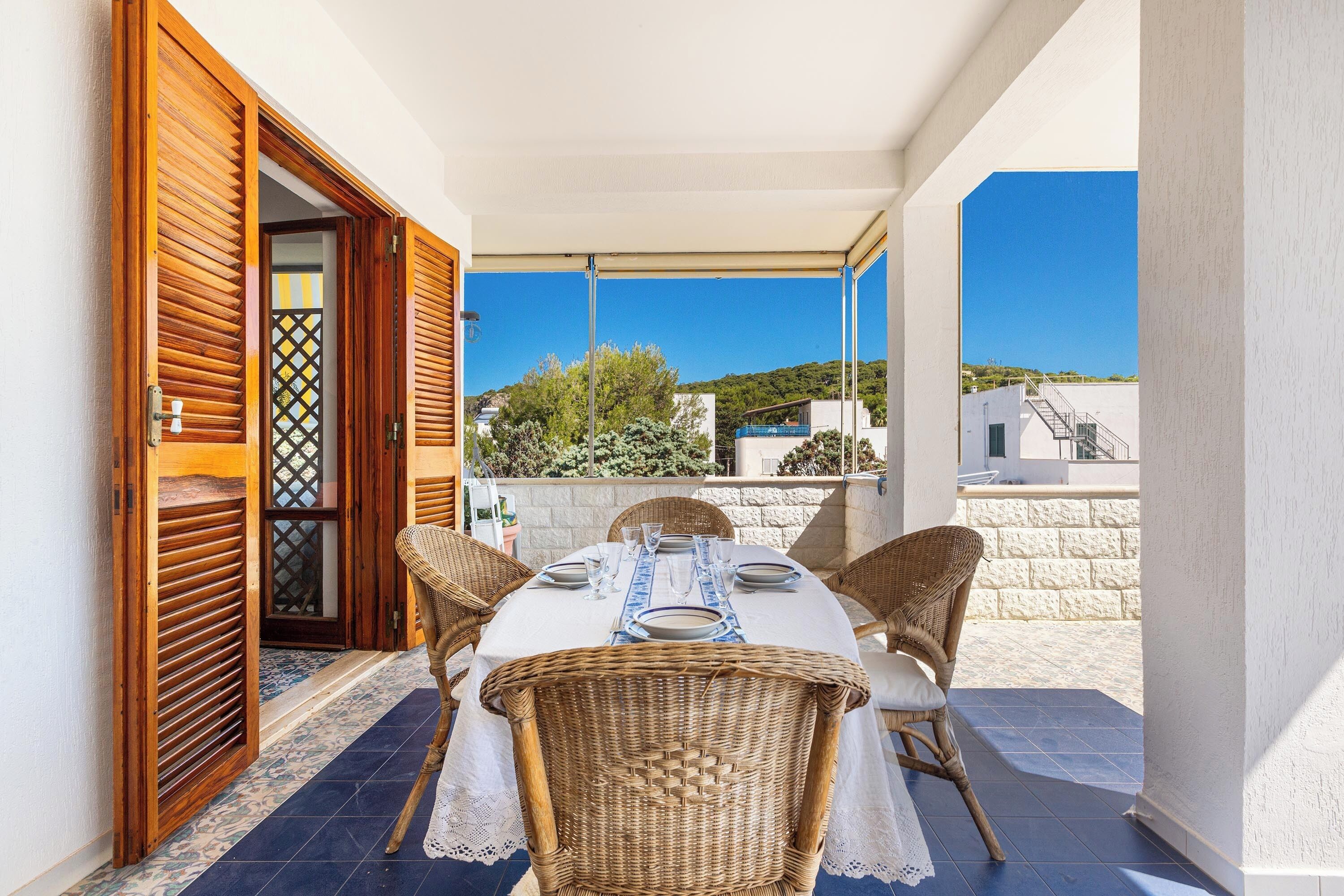 A balcony with a table set for two, wicker chairs, and a view of the surrounding landscape.