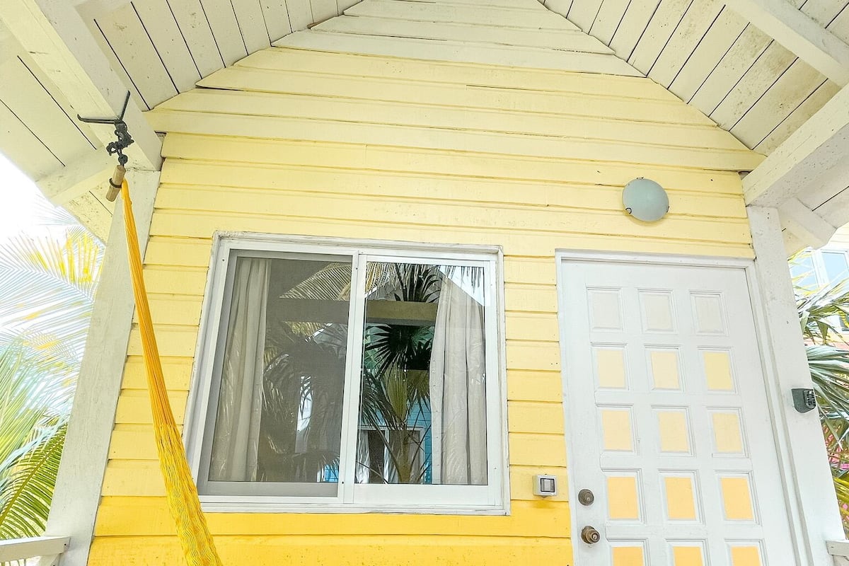 Um edifício amarelo com porta e janela brancas, uma rede e um teto com vigas de madeira.