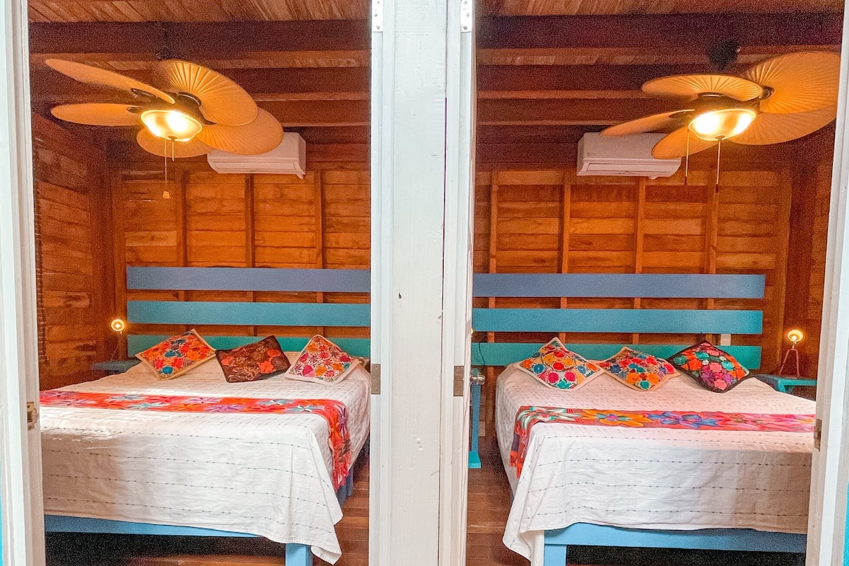 Reflexo num espelho de um quarto de madeira com duas camas, cada uma com colcha branca e almofadas coloridas, e um ventilador de teto.