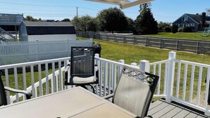 Een overdekt terras met een tafel en stoelen, omringd door witte leuningen, met uitzicht op een gazon met een hek en huizen op de achtergrond.