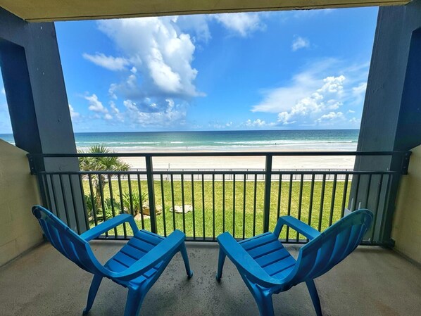 A balcony with two blue plastic chairs, a view of the beach, and a clear sky.