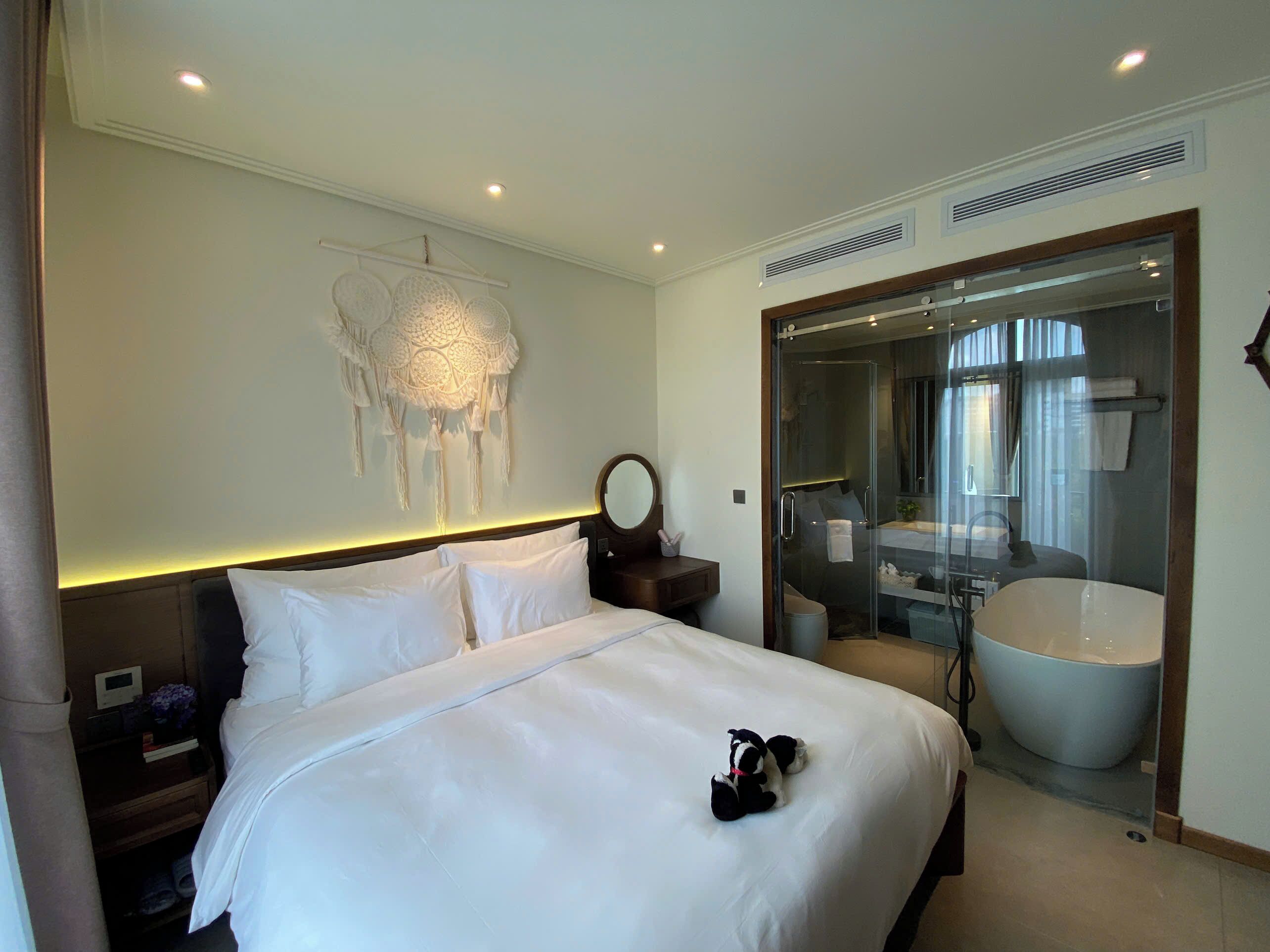 Une chambre d’hôtel avec un grand lit, une table de chevet, un attrape-rêves fixé au mur, une douche en verre et une salle de bain avec une baignoire et un lavabo.