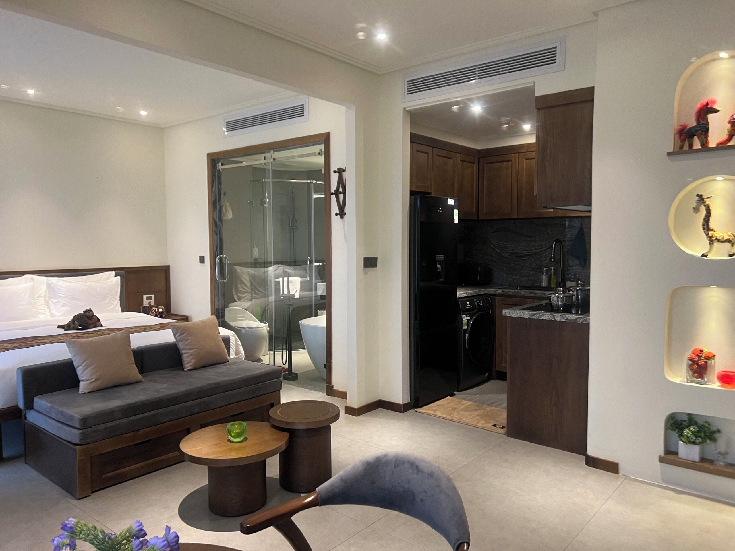 Une chambre d’hôtel moderne équipée d’un canapé, d’un fauteuil, d’une petite table, d’une douche en verre, d’un coin cuisine avec un réfrigérateur et d’un évier.