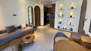 A modern living room with a sofa, a coffee table, and a chair. There is a kitchen in the background with a sink and cabinets. - JB Serviced Apartment Phu Quoc (Phu Quoc)