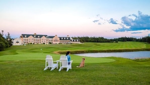 Campo de golf con club house al fondo, un estanque y dos sillas con un perro.