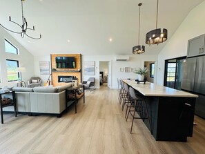 A modern kitchen with a central island, a dining area with bar stools, and a living room with a sofa and a fireplace.