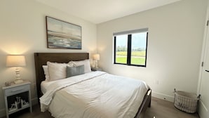 Un dormitorio con cama, una mesita de noche, una lámpara, una ventana con vistas y un cuadro en la pared.