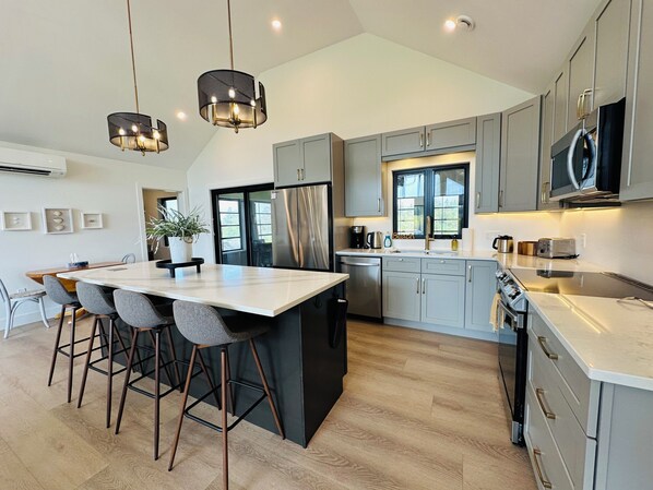 Een moderne keuken met een centraal eiland, krukken, roestvrijstalen apparatuur en hanglampen.