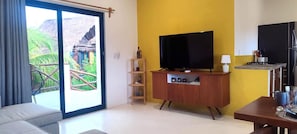 A living room with a TV, a sofa, a wooden cabinet, and a balcony with a view of a house and greenery.