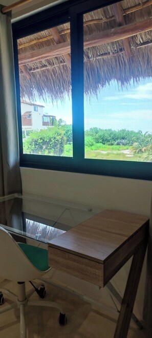 A room with a large window, a wooden desk, and a thatched roof visible through the window.